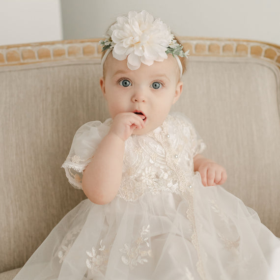 A baby with bright blue eyes sits on a beige sofa, wearing the Kristina Christening Gown & Bonnet, looking surprised with one finger near her mouth.