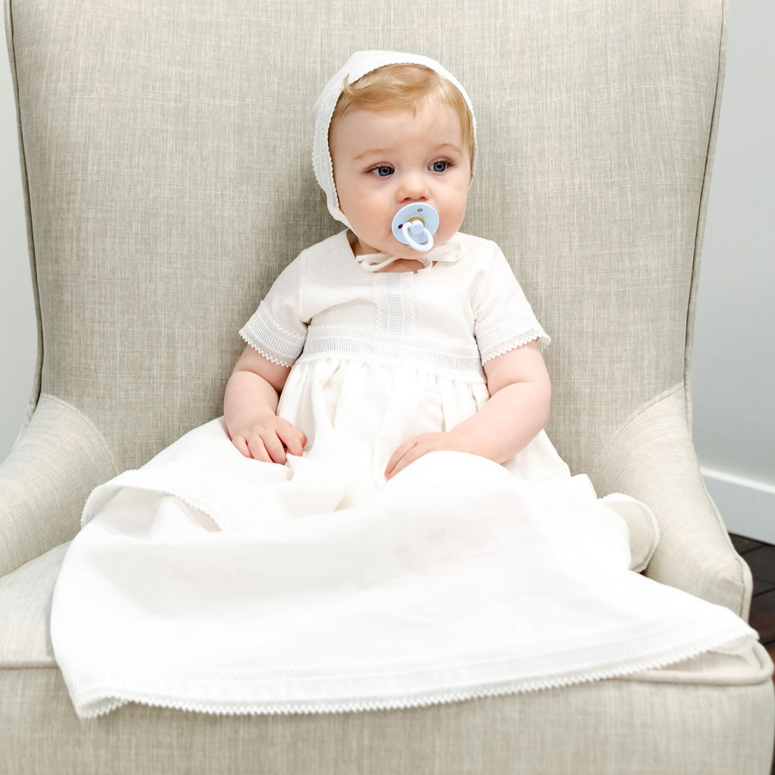 Baby boy dressed in a linen baptism gown sitting in chair with pacifier in mouth. 