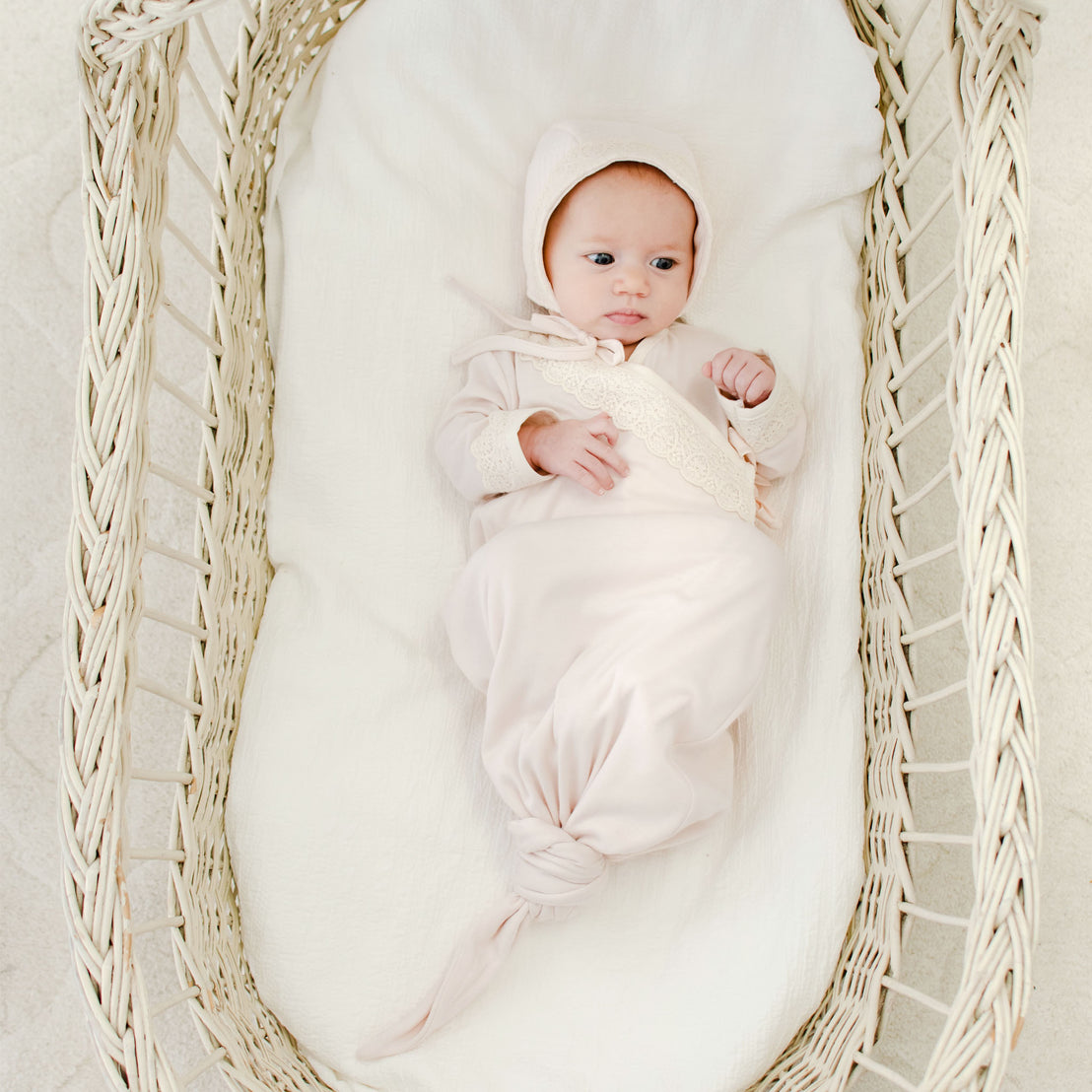 A newborn dressed in an Evelyn Knot Gown with matching Evelyn Quilt Bonnet, lying in a woven basket, looking upwards with a calm expression.