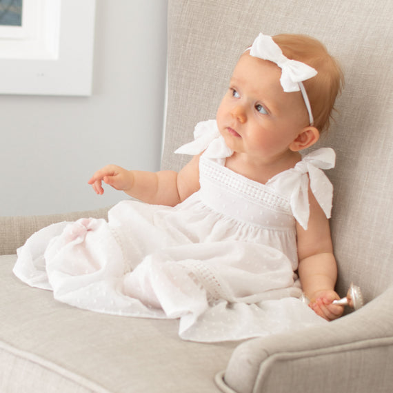 A newborn girl in the Mila Cotton Gown with ribbon details on the shoulders and the matching Mila Bow Headband on her head sits on a grey chair, looking to the side with a curious expression.