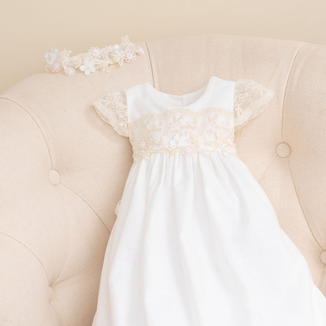 A Jessica Linen Gown with lace detailing displayed on a soft beige vintage sofa, complemented by the Jessica Beaded Flower Headband, a floral detailed lace headband, placed above it.