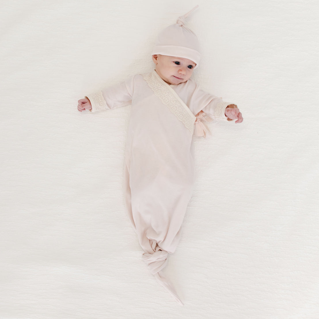 A newborn dressed in an Evelyn Knot Gown with the matching Ava Pima Knot Cap lies comfortably on a soft white blanket.