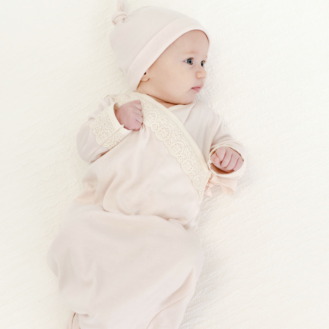 A newborn baby wearing the Evelyn Knot Gown and matching Ava Pima Knot Cap, looking off to the side while lying on a cream-colored background. The outfit is adorned with delicate lace detailing.