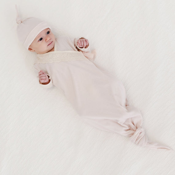 A newborn wearing the Evelyn Knot Gown and matching Ava Pima Knot Cap lies on a soft white background, looking upwards with a slight smile.