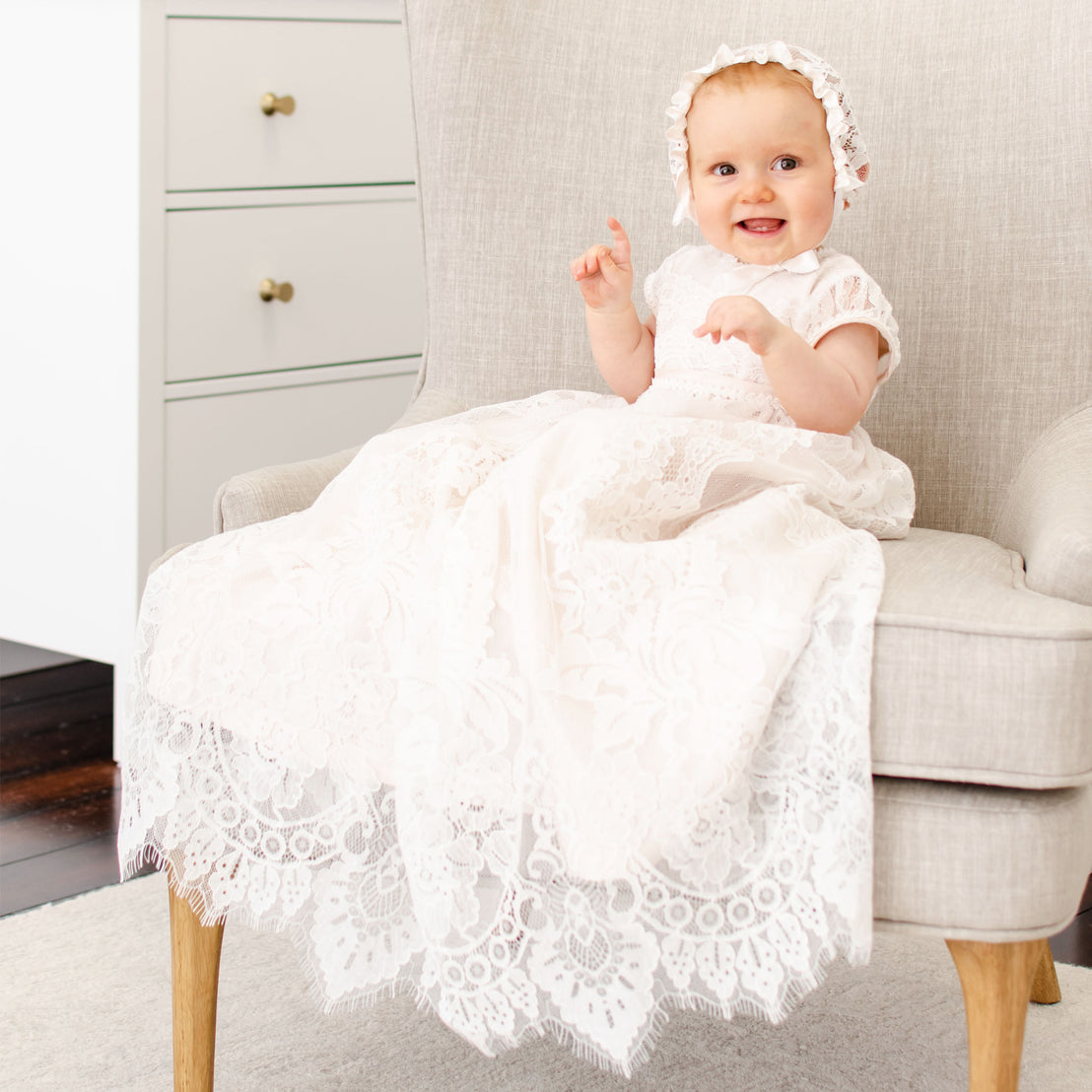 Baby girl sitting on a chair and wearing the pink Victoria Puff Sleeve Christening Gown & Bonnet. The gown is made from a pink silk Dupioni lining under an embroidered ivory floral lace overlay.
