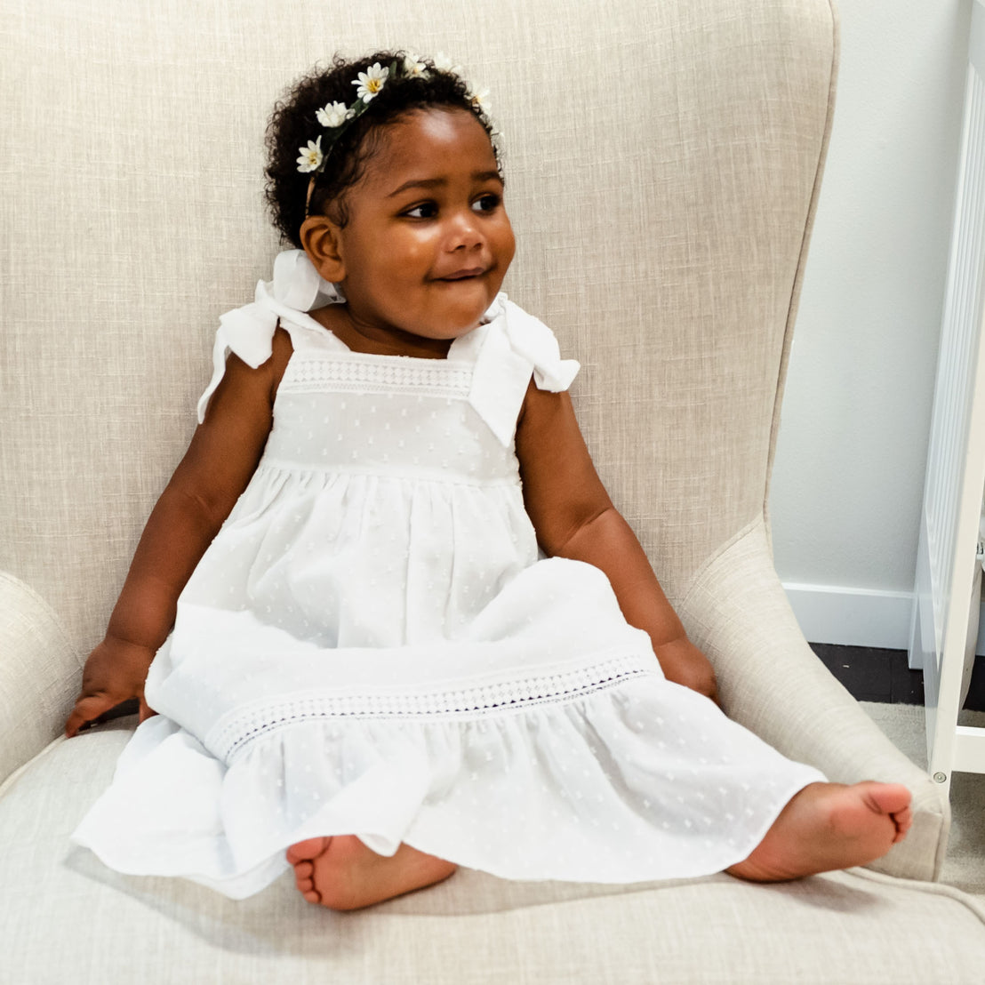 A toddler girl with a floral headband sits on a grey sofa, wearing a Mila Cotton Gown and looking off to the side with a subtle smile. 