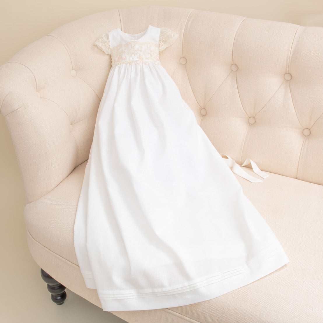 A white, elegant Jessica Linen Gown with vintage lace detailing on the bodice hangs on an upholstered beige tufted armchair in a soft, well-lit room.