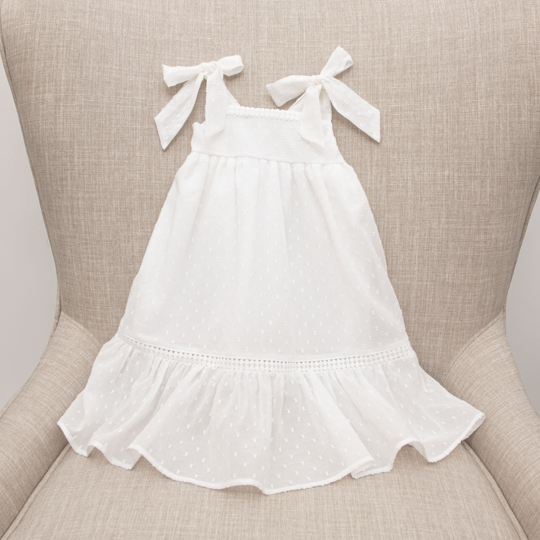 The Mila Cotton Gown with tie straps on the shoulders and a ruffle hem displayed on a beige upholstered chair. The gown features delicate lace detailing near the hemline.