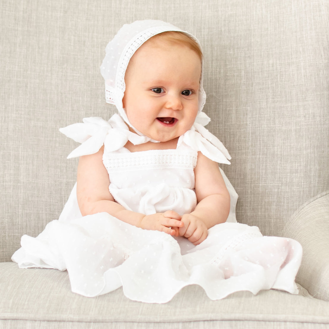 A joyful baby girl wearing the Mila Cotton Gown & Bonnet sits on a grey sofa, smiling and looking to the side with a playful expression.