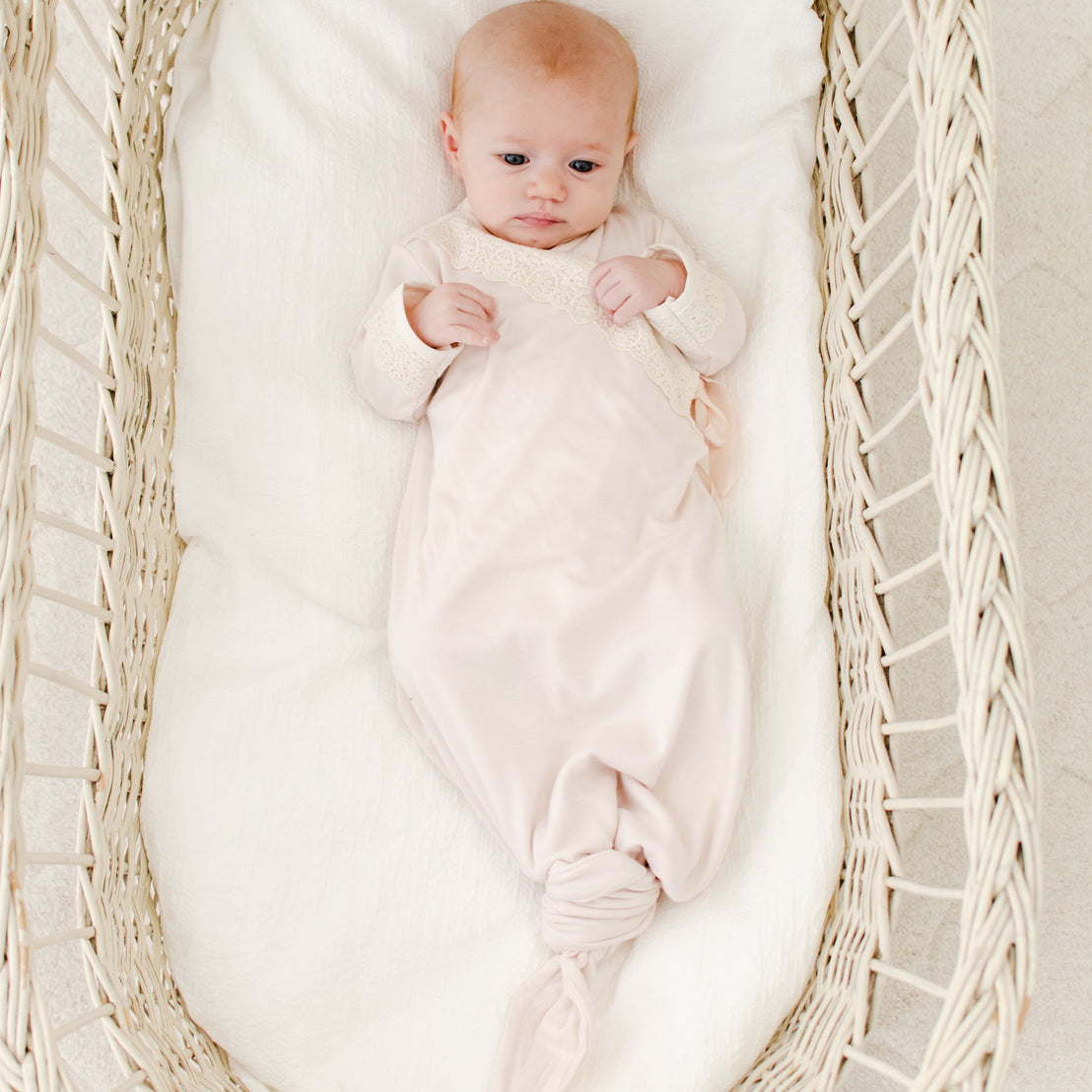 A newborn wearing the Evelyn Knot Gown lies in a white woven bassinet, looking upward with a calm expression. The soft colors and light create a serene atmosphere.