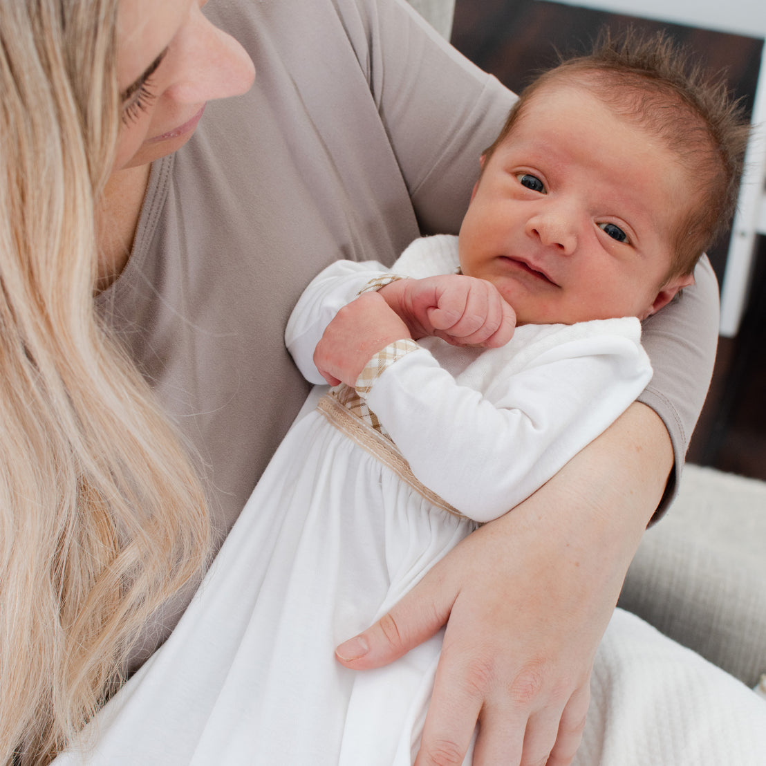 A mother gently holds her newborn baby, who is looking directly at the camera. The baby is dressed in a white Dylan Layette outfit with a bow, and the mother is smiling lovingly down at them.