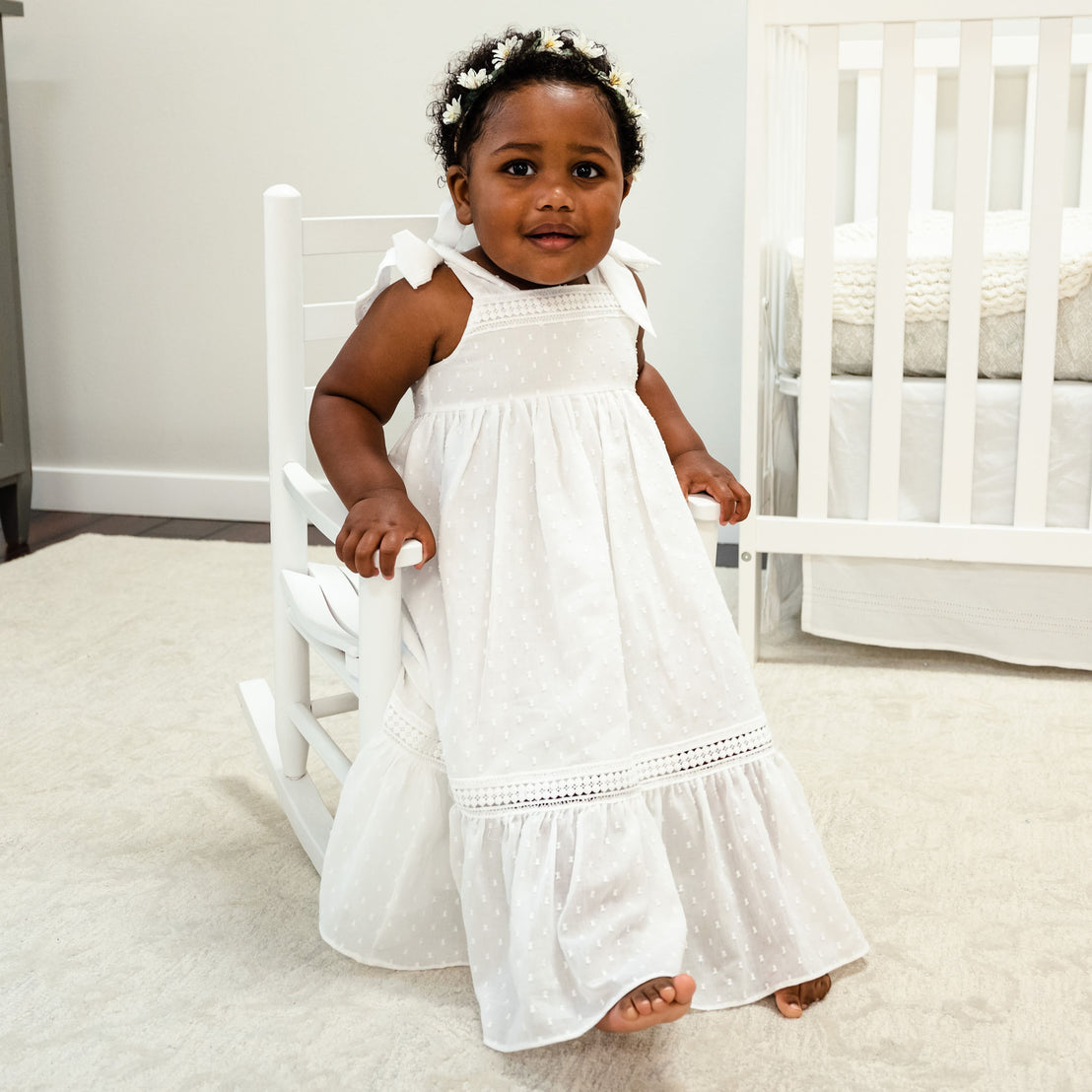A toddler girl with a floral headband sits on a white rocking chair, wearing a Mila Cotton Gown and looking at the camera with a subtle smile. In the background to the left is a white crib.