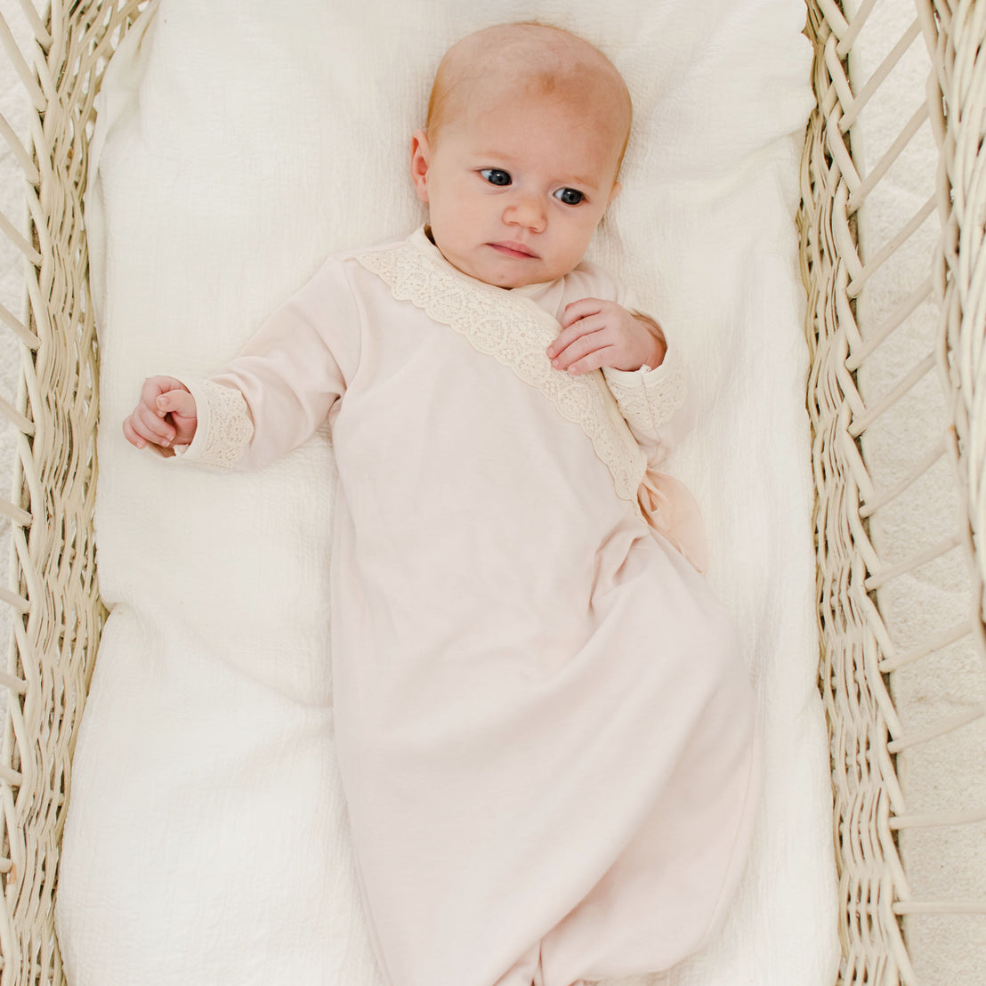 A newborn wearing the Evelyn Knot Gown with lace detailing lies in a white wicker basket, looking up with a calm expression.