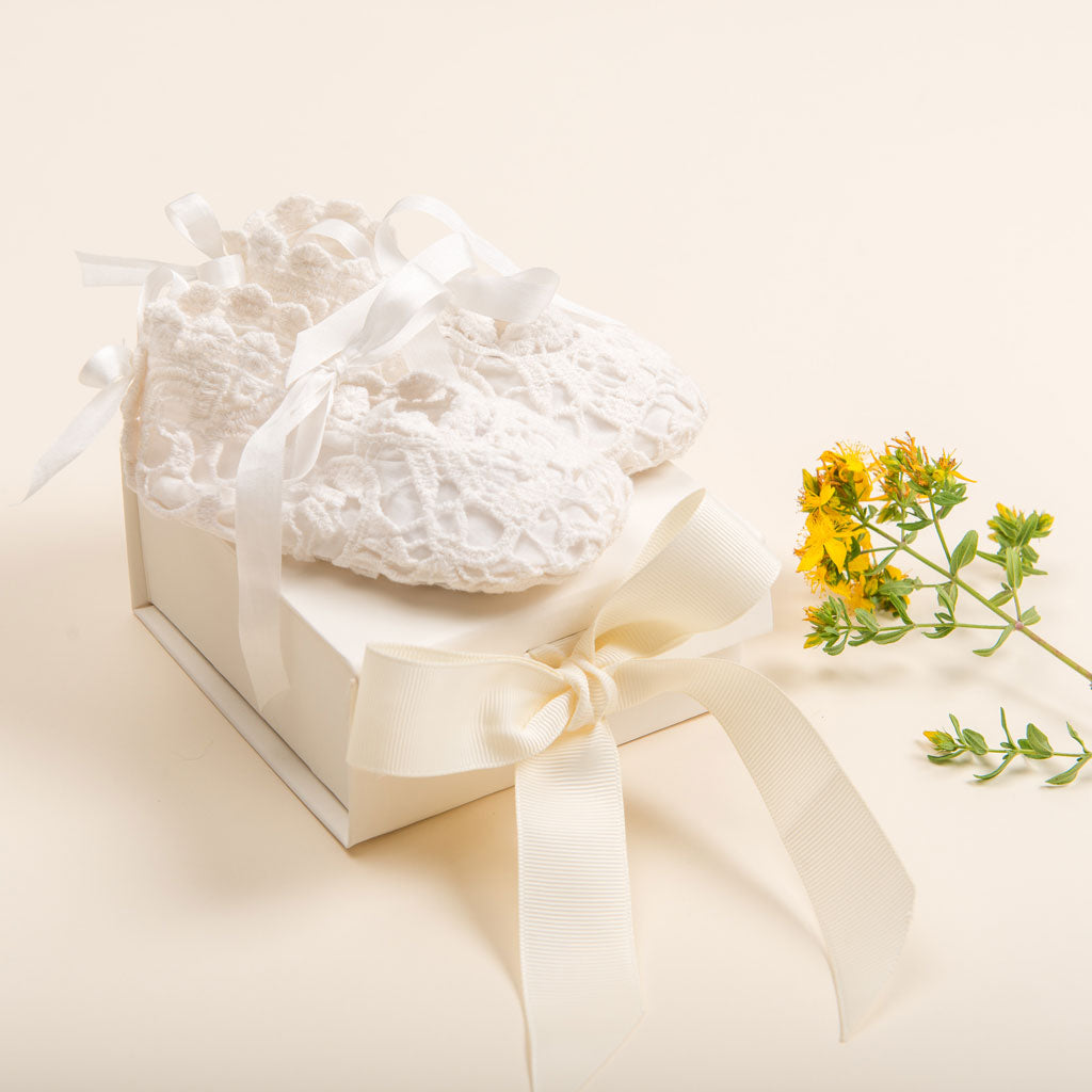 A pair of white lace baby booties on top of a Baby Beau & Belle Ivory Gift Box tied with a cream ribbon, accompanied by small yellow flowers on a light beige background.