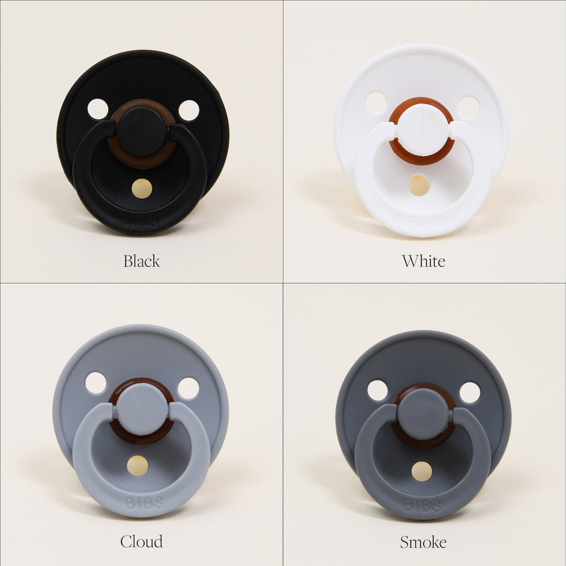 Four Bibs Pacifiers in different colors labeled as black, white, cloud, and smoke, displayed against a light background. Each natural rubber baby pacifier features a ring handle and three ventilation holes.