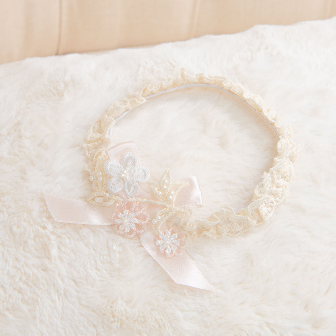 The Jessica Headband, a delicate floral lace headband with soft pink ribbon accents, rests on a soft white background.
