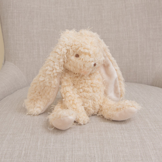 A fluffy, cream-colored Harey Bunny with long ears and droopy eyes dressed in a boutique outfit sitting on a beige sofa, giving a soft and cozy appearance.