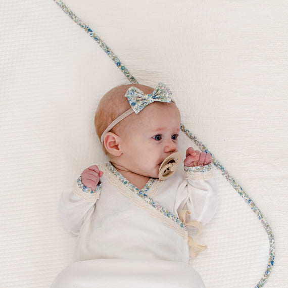 A baby wearing a light, vintage-inspired dress and a blue Petite Fleur Headband, holding a tan pacifier, lies against a white background, framed by an heirloom blue and white braided rope.