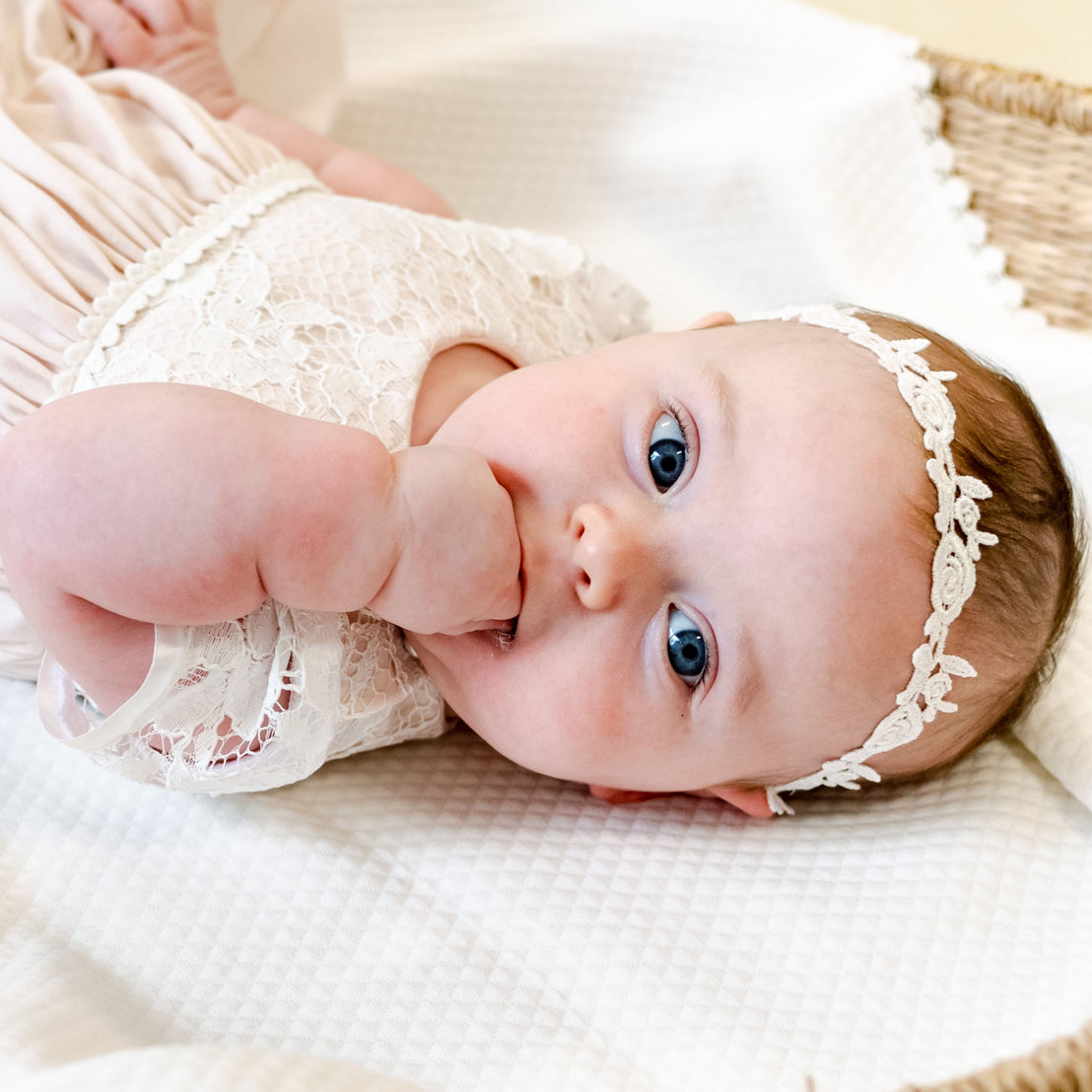 A baby with blue eyes lies on a cream blanket, sucking her thumb while gazing at the camera during a traditional baptism wearing the Rose Lace Headband.