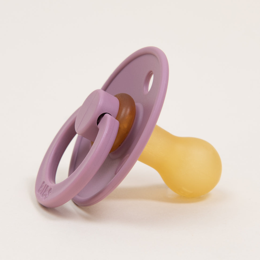 A purple Bibs Pacifier in Heather lying on a white surface. The pacifier has a purple handle and rim, with a beige nipple.