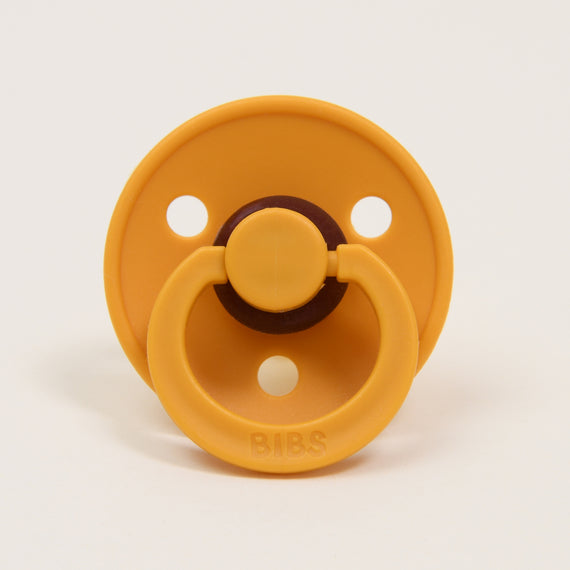 A honey bee natural rubber baby pacifier with a circular shield and two ventilation holes, featuring a dark brown button handle in the center. The brand "bibs" is embossed at the top.