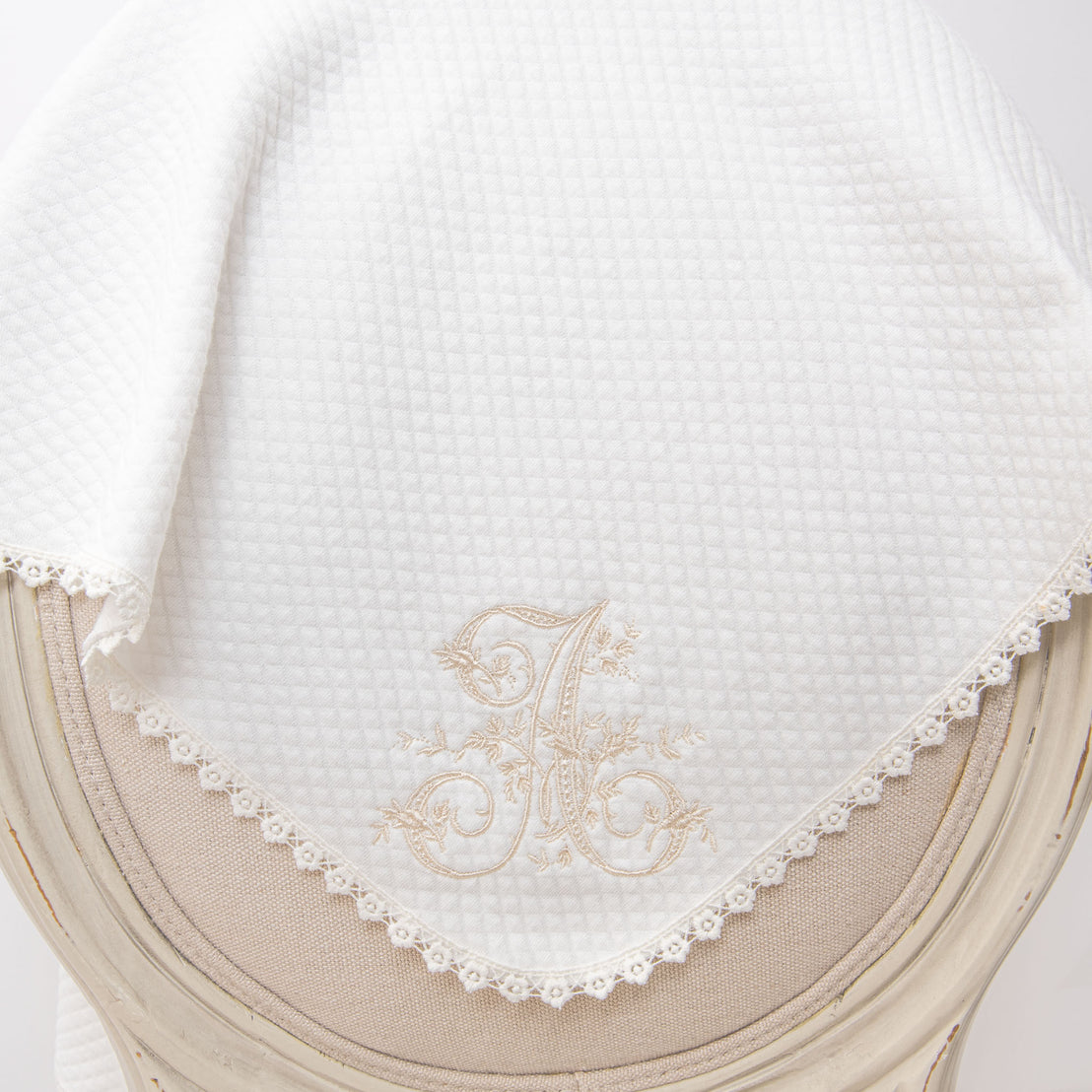 Elegant white baby blankets with initial embroidery personalization 'a' and delicate lace trim, draped over the edge of a soft beige cushioned chair.