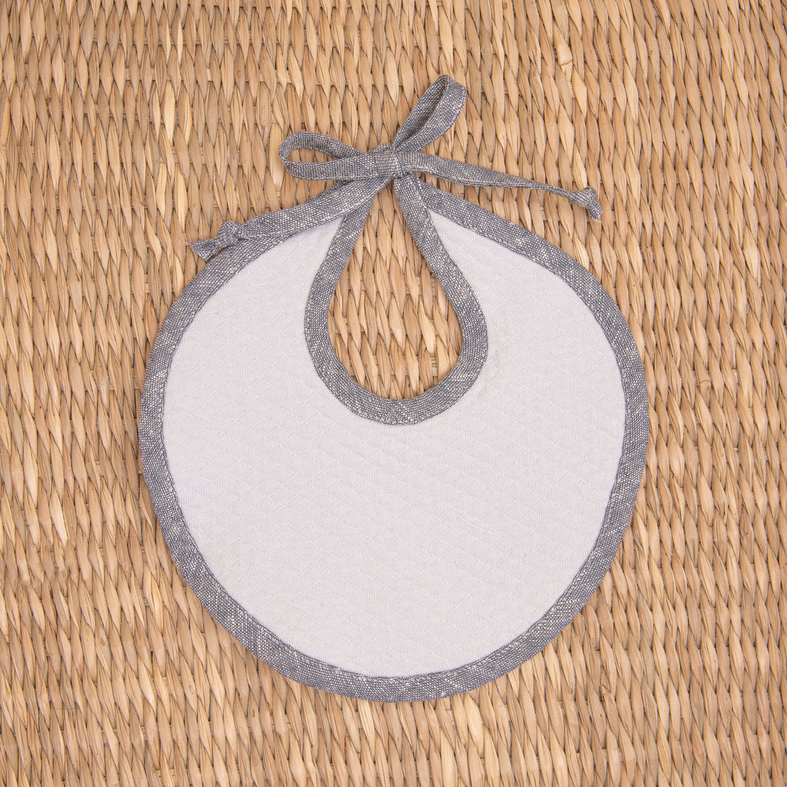 Flat lay photo of the blue Silas Linen Trim Bib. The bib is made from textured cotton in light grey