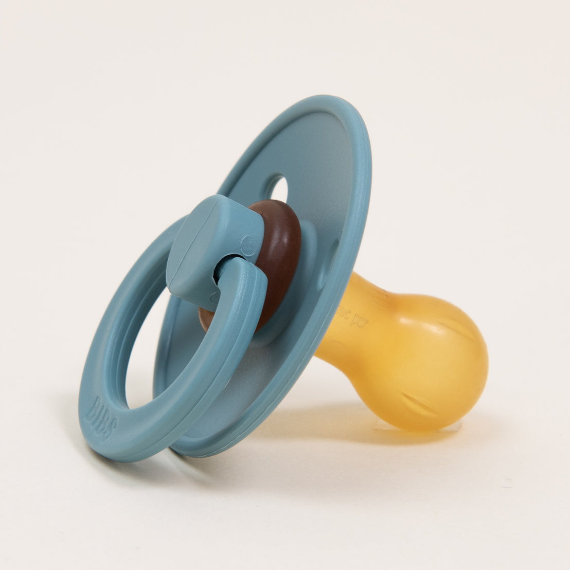 A baby's Bibs Pacifier in Island Sea, designed and manufactured in Denmark, with a teal ring, brown knob, and translucent yellow rubber nipple on a white background.