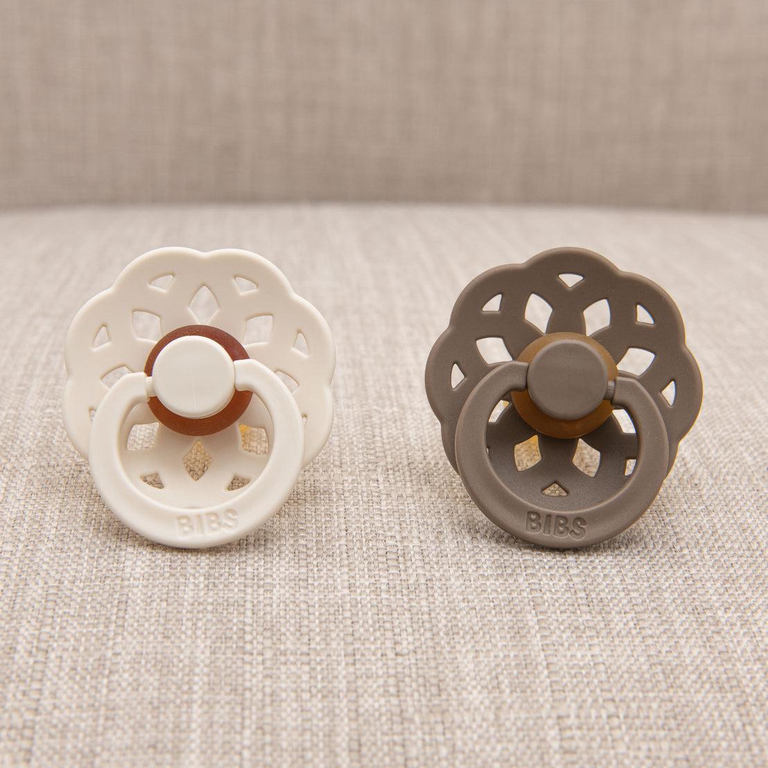 Two Madeline pacifiers, one ivory and one dark oak, rest on a textured gray fabric surface.