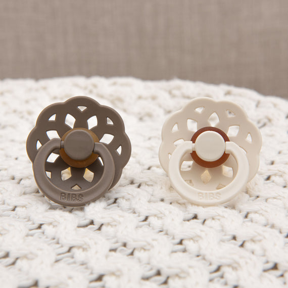 Two Madeline pacifiers, one dark oak and one ivory, on a white crocheted mat with a beige background, perfect for a baby gift.