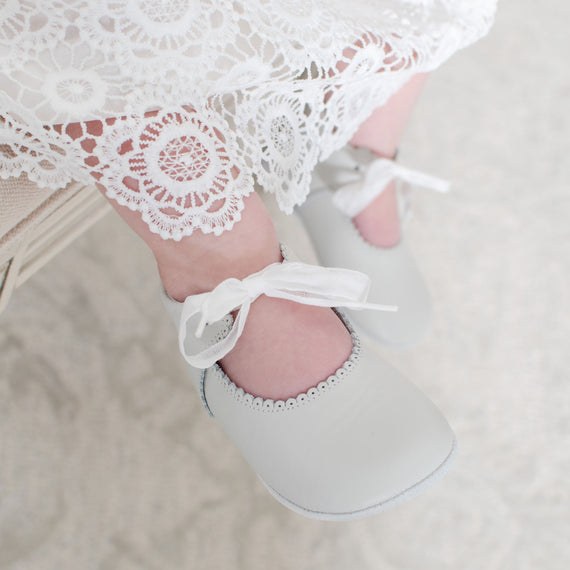 A close-up of a child's feet wearing Ivory Tie Mary Janes with white ribbons and scalloped edges, under a white lace dress. The background shows a soft, textured surface.