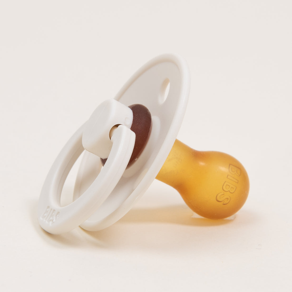 A close-up of a Bibs pacifier with a white and brown nipple and a transparent handle, set against a plain white background.