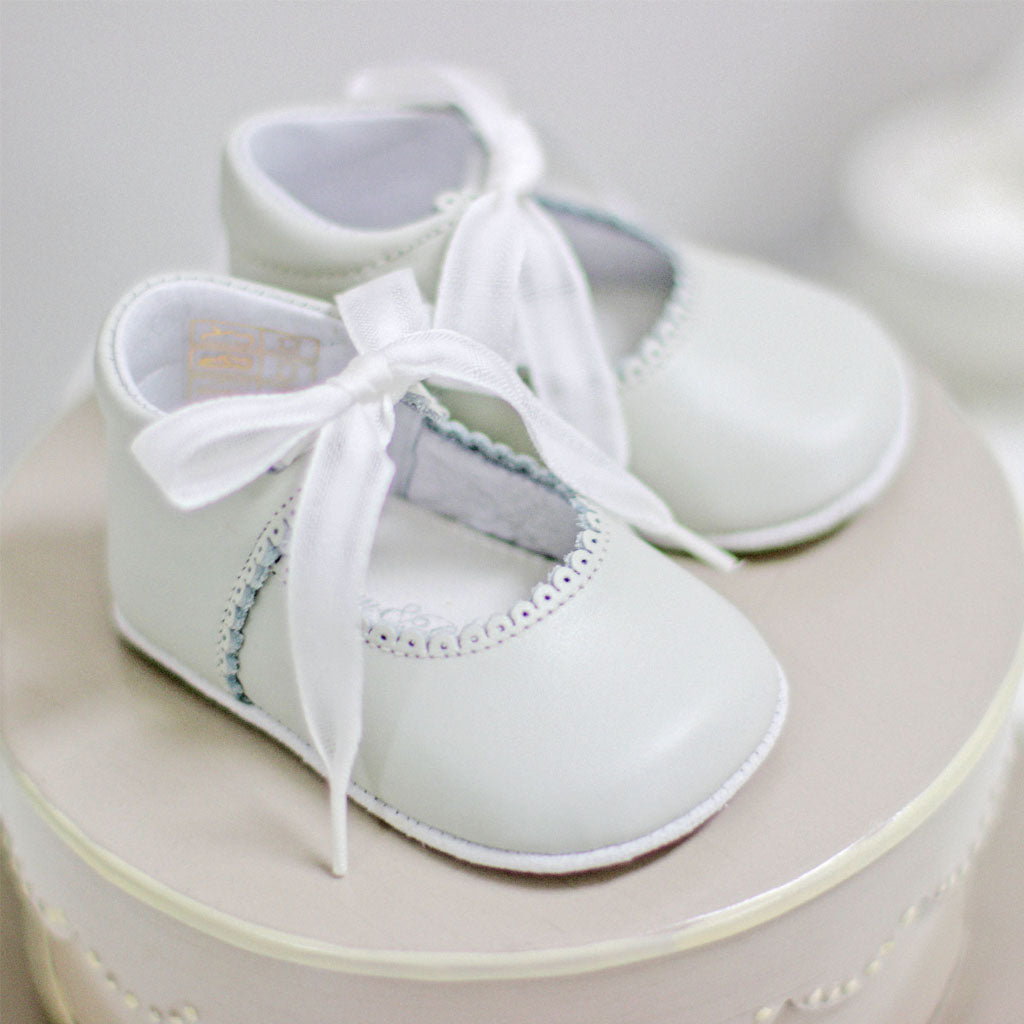 A pair of ivory tie Mary Janes with white ribbon laces, set atop a matching round cake, suggesting a celebration like a baby shower or a first birthday.