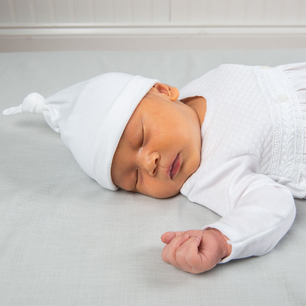 A newborn baby peacefully sleeping, dressed in a white knitted layette and Dylan Knot Cap, lying on a soft grey blanket.