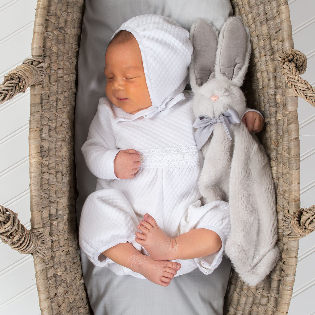 A newborn baby sleeps in a cradle. He is wearing the Elijah Newborn Romper made from a plush white quilted cotton, featuring white Venice lace at the bodice and white linen trim at the cuffs and neck