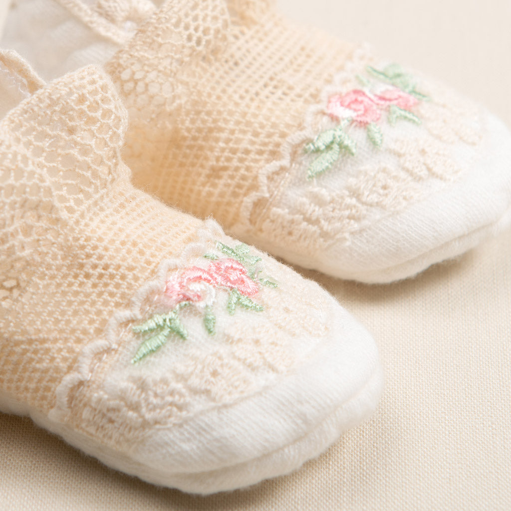 Close-up of vintage white knitted Clementine Booties featuring delicate pink and green floral embroidery on a soft beige background, perfect for baptism.