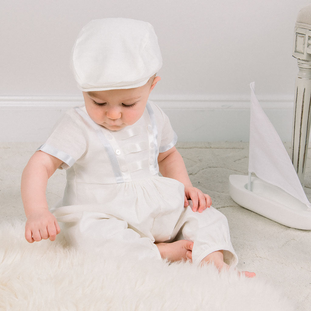 A baby dressed the Owen Romper and Newsboy Cap sits on a fluffy rug, looking curiously at the floor, with a white sailboat model nearby.