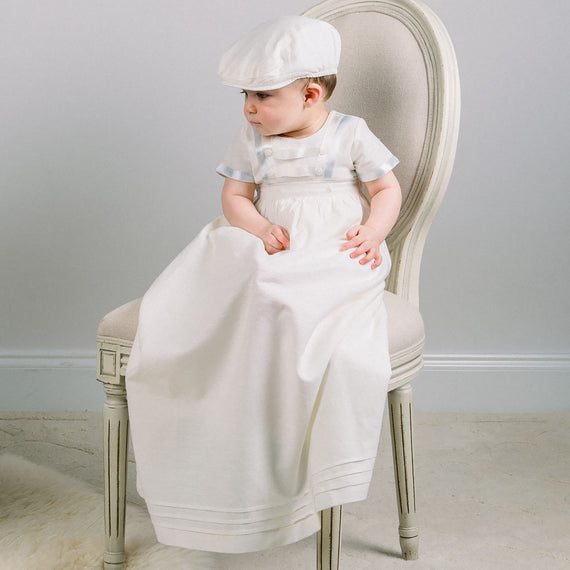 A toddler in an Owen Linen Convertible Skirt sits on an elegant chair, draped in a long white cloth, looking thoughtfully to the side. The background is neutral, emphasizing the child's vintage attire and pose.