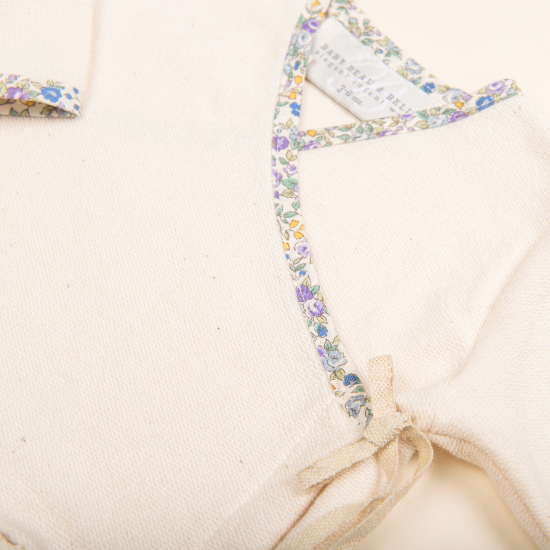 Close-up of a Petite Fleur Wrap Top & Pants, an upscale, handcrafted beige textile with a delicate floral trim and a tag reading "made in Italy," featuring a tied fabric bow at the seam.