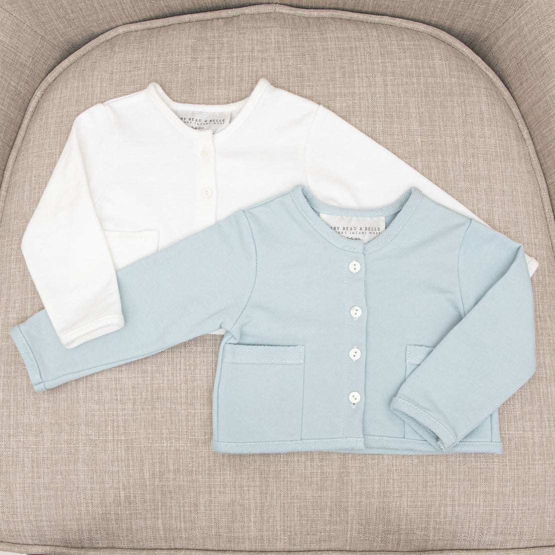 Two Isla French Terry Jackets, one white and one light blue, each featuring heirloom quality. They are displayed on a beige textured cushion. The blue jacket has buttons and a pocket detail.