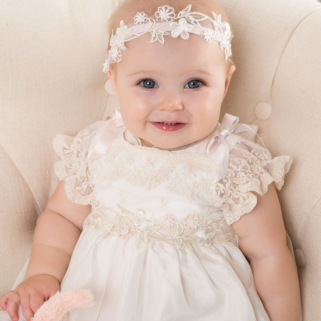 A smiling baby sitting on a beige sofa and wearing the Jessica Beaded Flower Headband along with the Jessica Linen Gown.