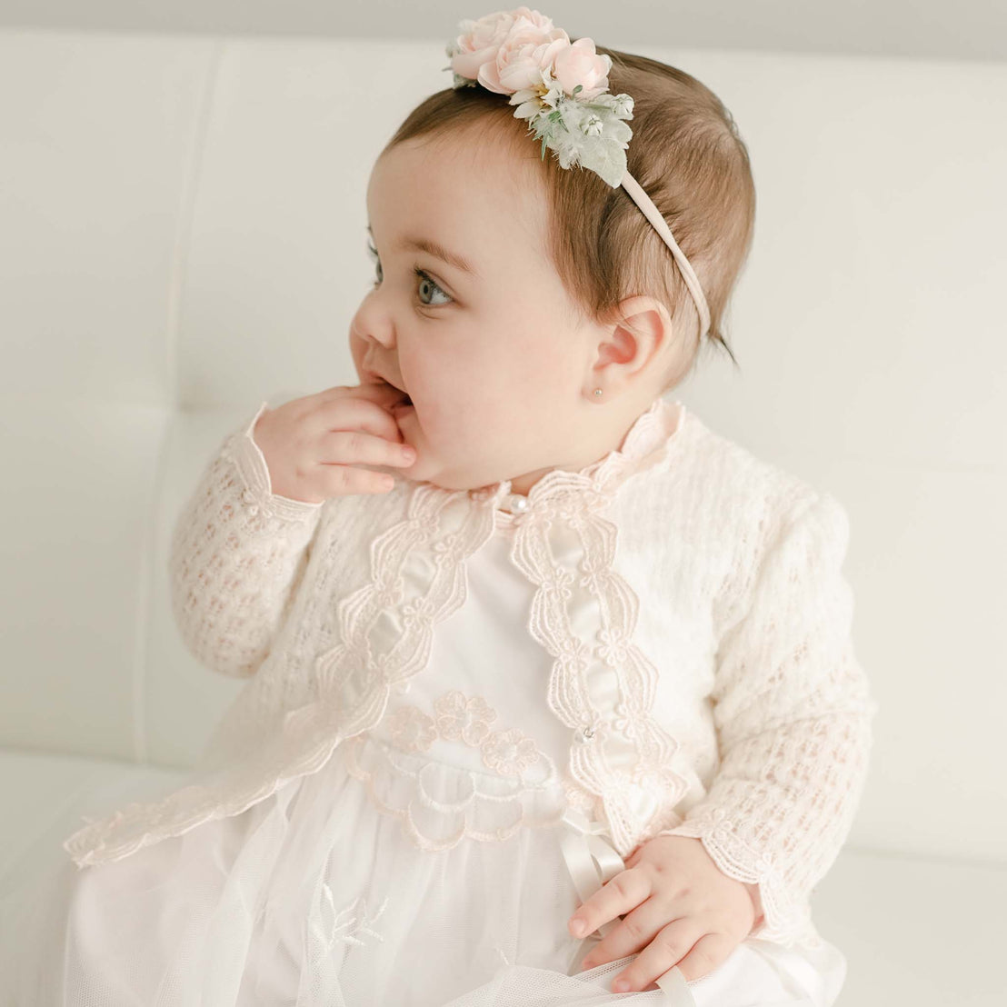 A baby girl in a Joli Knit Sweater & Bonnet, looking to the side with her hand near her mouth, dressed for a baptism against a soft, light background.