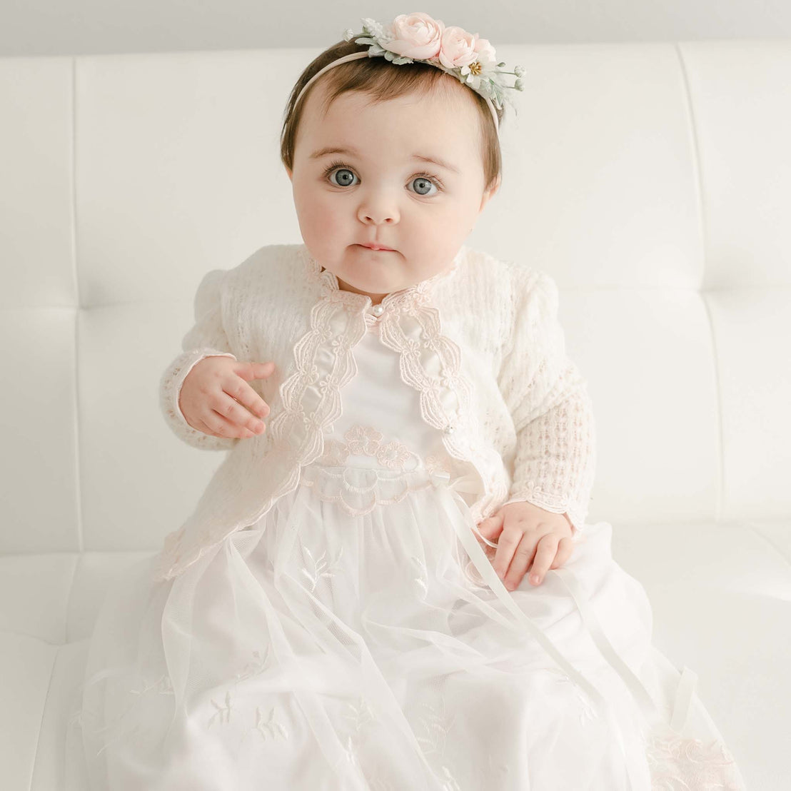 A baby girl with blue eyes wearing a Joli Knit Sweater & Bonnet sits on an upscale white sofa, looking surprised.