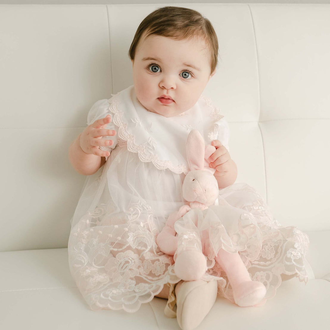 A baby with big blue eyes, wearing a delicate white lace dress, sits on a sofa holding a Joli Silly Bunny Buddy Pacifier Holder.