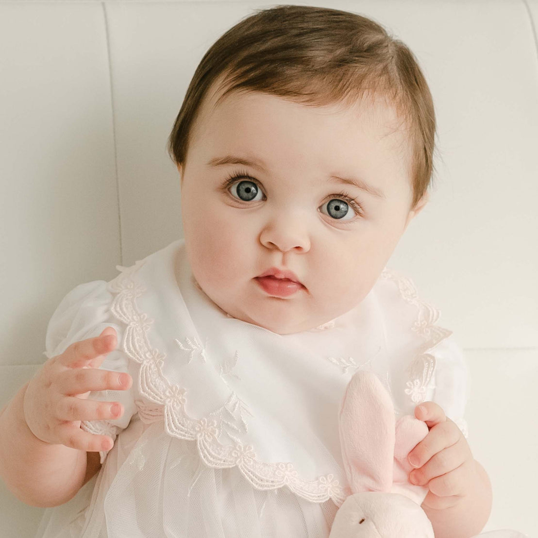 A baby with blue eyes and brown hair poses in a vintage-inspired white Joli Bib, holding a small pink bunny for her baptism. The child appears curious and attentive.