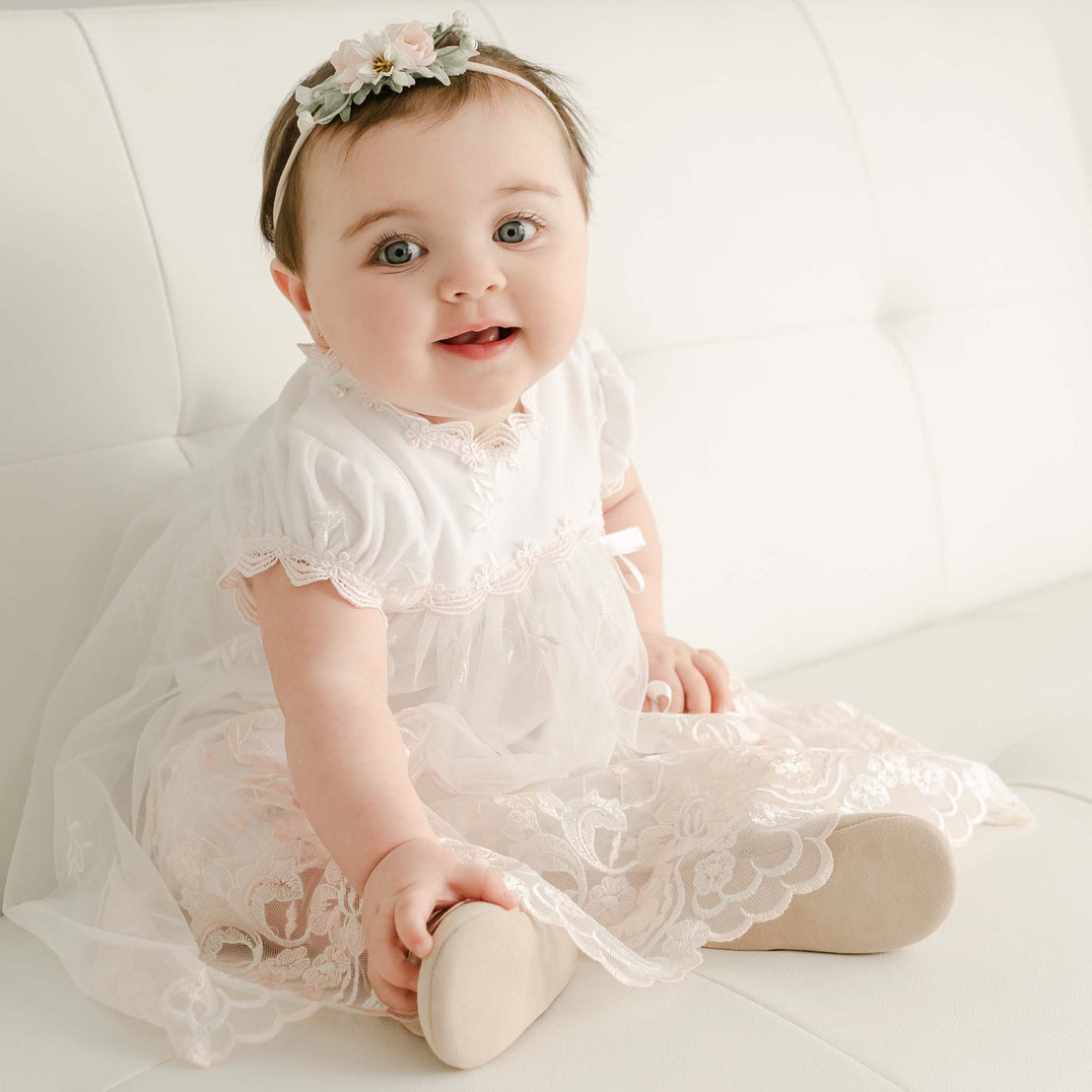 A baby girl in a Joli Romper Dress and headband with floral detail sits smiling on an upscale white sofa, looking directly at the camera.