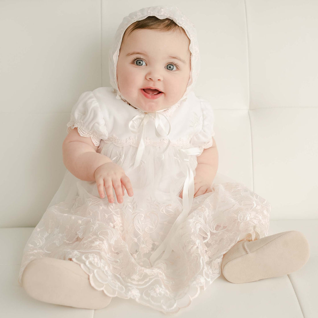 A joyful baby with blue eyes, wearing a Joli Romper Dress and vintage-inspired matching bonnet, sits on a white sofa, looking up with a wide smile.
