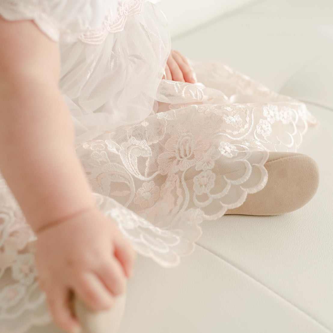 A close-up view of a baby in a Joli Romper Dress for baptism, focusing on the detailed fabric and the baby's hand gently touching the dress.