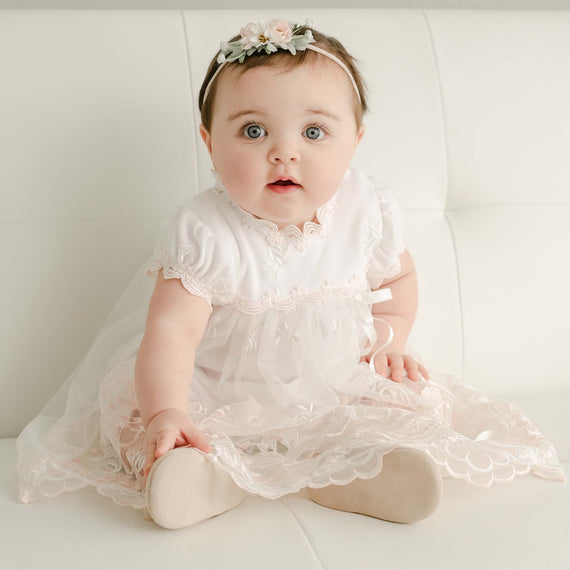 A baby girl sitting on a white sofa, wearing a Joli Romper Dress and a floral headband, with big, curious blue eyes looking directly at the camera.