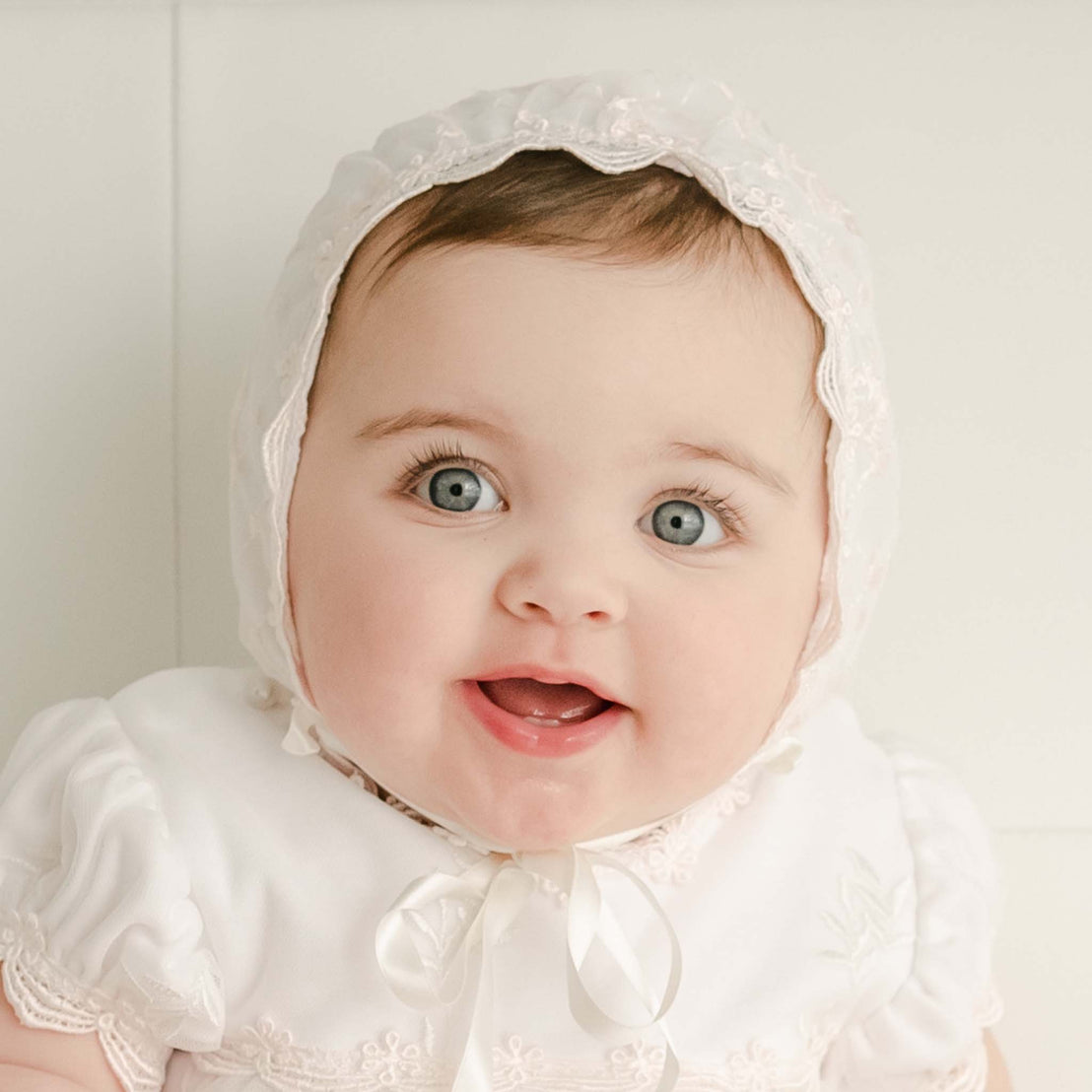 A cheerful baby dressed in a vintage-inspired, white baptism dress and Joli Fitted Bonnet with lace detailing, looking upwards with big blue eyes and a joyful expression.